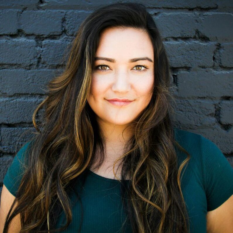 Smiling woman with long wavy hair against a black brick wall.